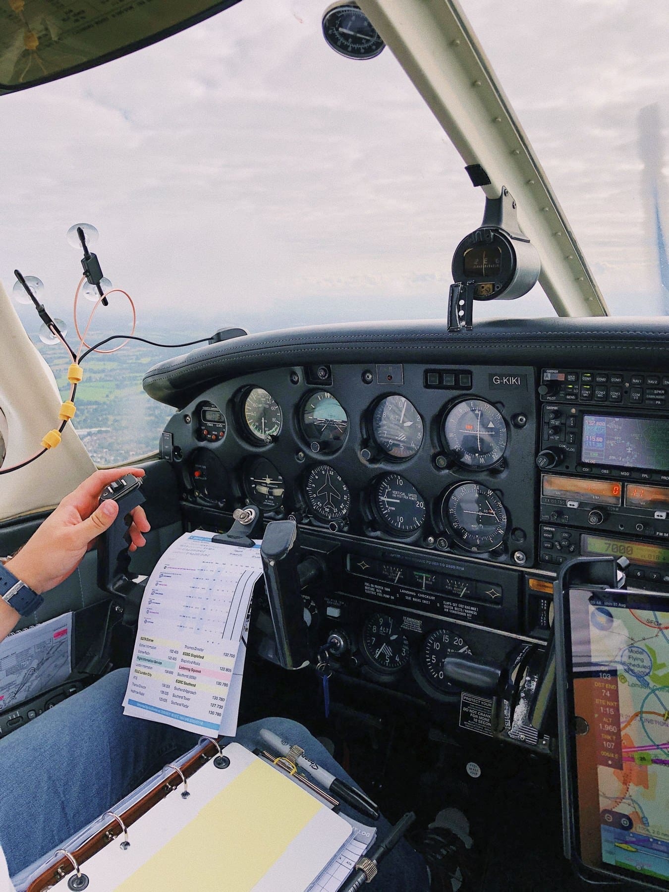 Pilot flying a light aircraft over the English countryside, with the cockpit instruments and navigation notes in view